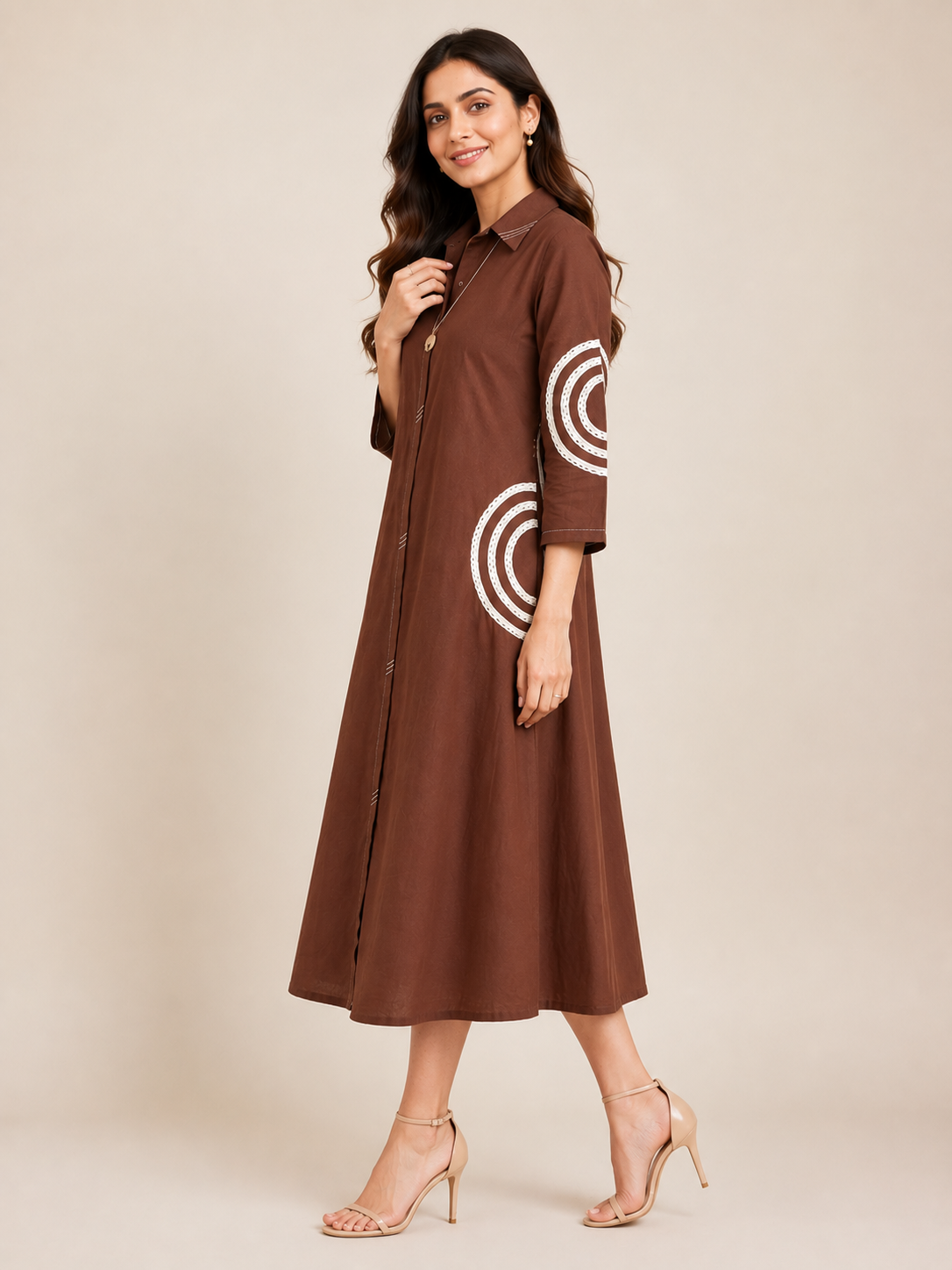 A woman modeling a brown midi shirt dress with white concentric circle patterns on the sleeves, paired with beige high-heeled sandals against a solid beige studio background.