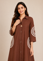 A woman wearing a brown button-up tunic dress with white concentric circle patterns on the sleeves and lower skirt, photographed against a plain beige studio background.