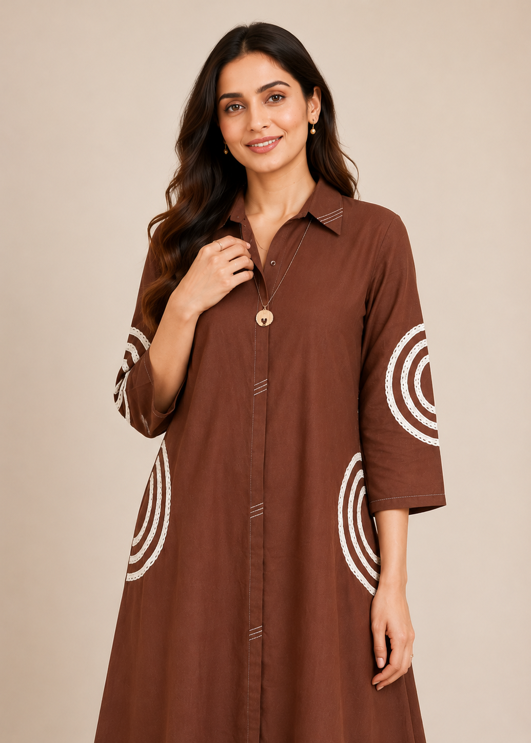 A woman wearing a brown button-up tunic dress with white concentric circle patterns on the sleeves and lower skirt, photographed against a plain beige studio background.