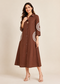 A woman modeling a brown midi shirt dress with three-quarter sleeves and white concentric circle patterns on the sleeves and pockets, set against a solid beige studio background.