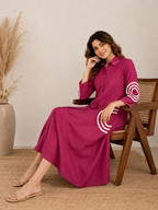 A woman wearing a magenta long-sleeve collared dress with white circular stripe details on the sleeves and hem, sitting on a wooden chair in a neutral indoor setting.