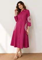 A woman modeling a magenta pink long-sleeve midi dress with white circular concentric pattern details on the sleeves and skirt, standing against a white wall.