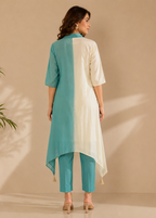 A woman stands with her back to the camera wearing a two-tone tunic and trouset set. The tunic has a vertical color-blocked design with teal green on the left and off-white on the right, paired with teal green cropped trousers.