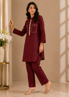 A woman models a matching deep maroon two-piece kurta and trouser set in a warm beige interior. The kurta features a high collar, a decorative gold-toned embroidered vertical line down the front, and two front patch pockets with button details.