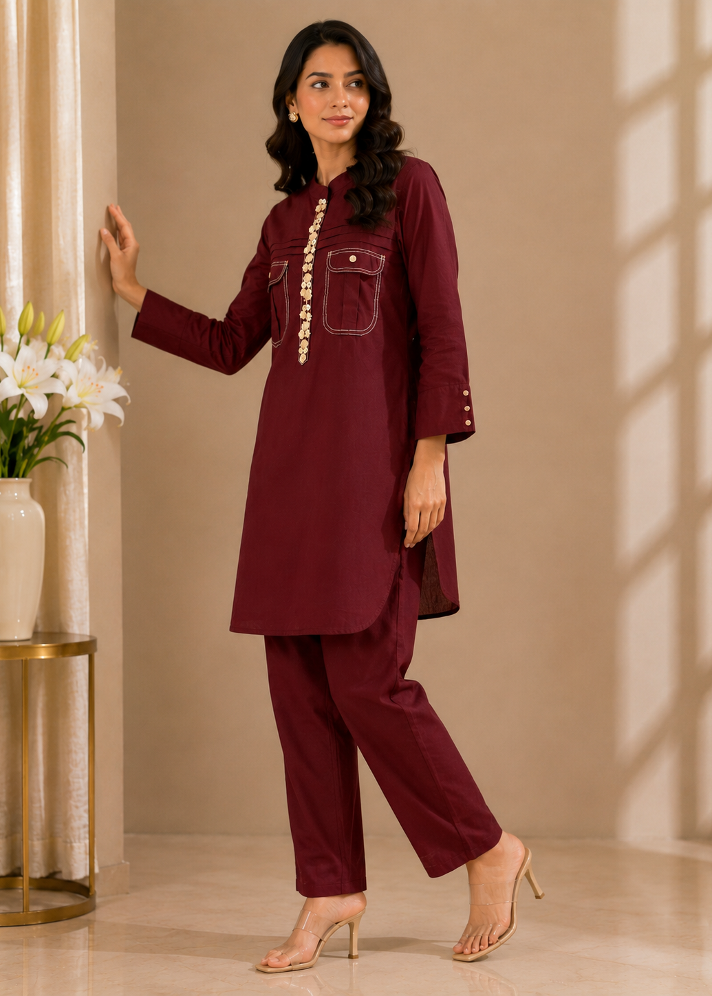 A woman models a matching deep maroon two-piece kurta and trouser set in a warm beige interior. The kurta features a high collar, a decorative gold-toned embroidered vertical line down the front, and two front patch pockets with button details.
