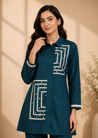 A woman modeling a dark teal long-sleeve kurta-style top with white geometric embroidery and matching pants, set against a warm beige background with soft shadows.