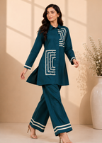A woman models a dark teal two-piece kurta set with white geometric embroidery and striped details, standing in a studio with a beige background and a large white vase.