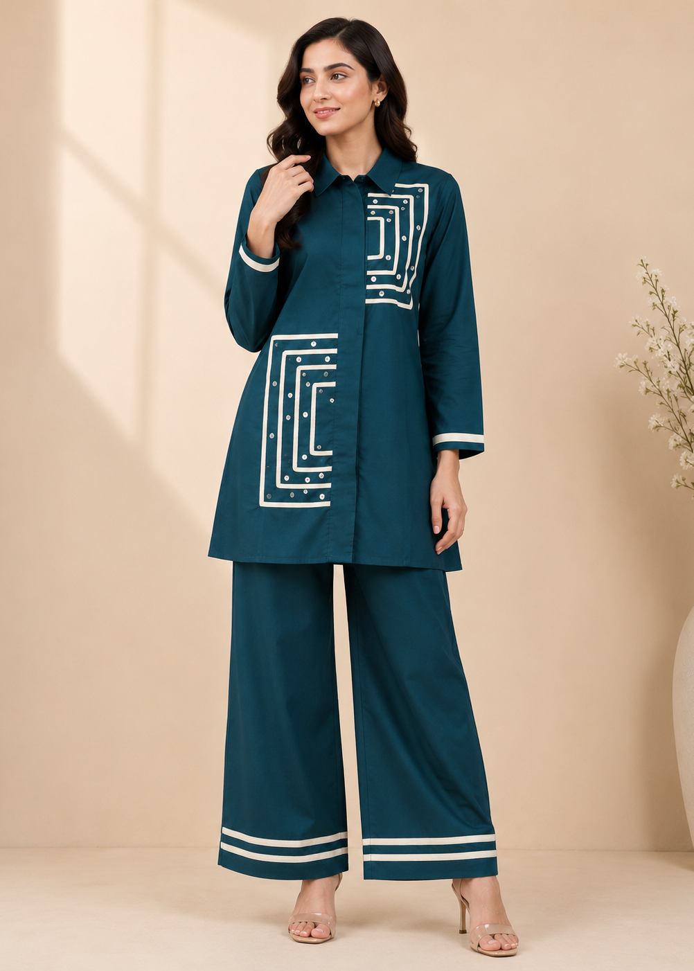 A woman modeling a dark teal two-piece kurta set with white geometric embroidery and striped details, standing against a beige studio backdrop.
