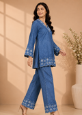 A woman models a blue denim two-piece set featuring a long-sleeve tunic with floral embroidery on the hem and cuffs, paired with matching wide-leg pants, set against a warm beige background with decorative shadows and a vase of flowers.