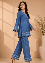 A woman models a blue denim two-piece set featuring a long-sleeve tunic with floral embroidery on the hem and cuffs, paired with matching wide-leg pants, set against a warm beige background with decorative shadows and a vase of flowers.