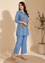A woman models a light blue two-piece kurta set with matching pants, featuring metallic gold embroidered details on the front. She is standing on a round woven rug against a beige wall with a plant in the background.