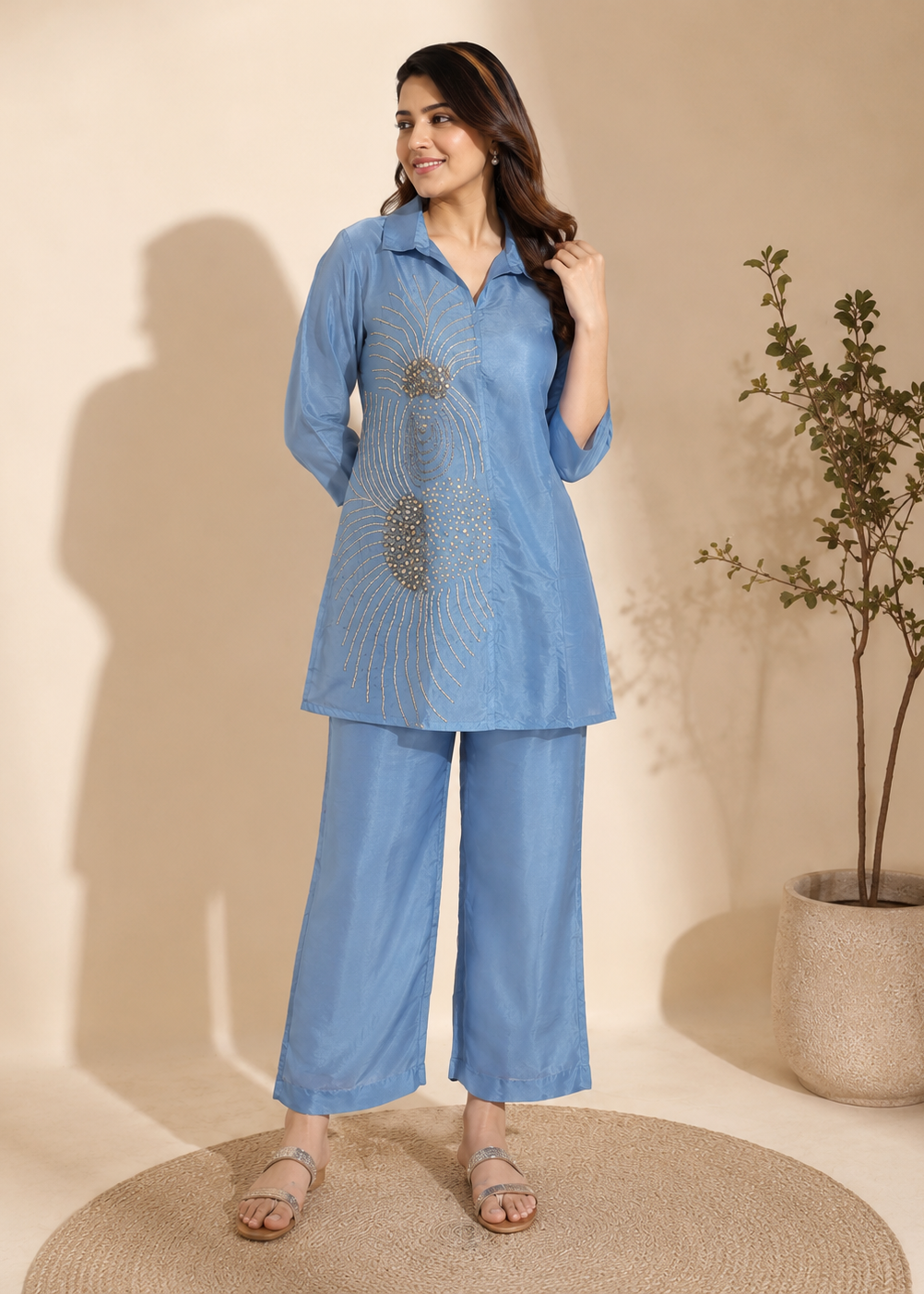 A woman models a light blue two-piece kurta set with matching wide-leg pants. The kurta features a button-up collar and a large, detailed gold and silver geometric embroidery on the front.