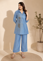 A woman models a light blue two-piece kurta set with matching wide-leg pants. The kurta features a button-up collar and a large, detailed gold and silver geometric embroidery on the front.