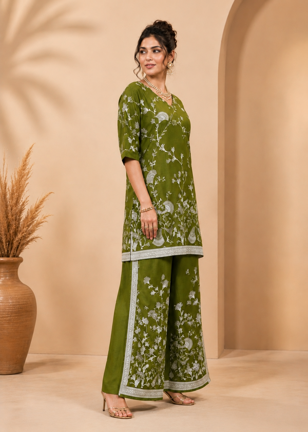 A woman models a three-piece olive green kurta set with white floral prints and light gray borders, standing in a warm beige studio setting with a potted plant and archway background.