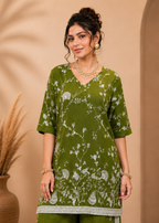 A woman modeling a green three-piece traditional outfit with white floral embroidery and silver border detailing, set against a warm beige background with a decorative vase.