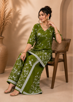 A woman seated on a wooden chair wearing an olive green traditional outfit with white floral print and silver border trim, posed in a warm beige interior with a large vase of dried grass in the background.