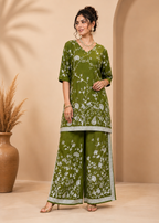 A woman models a three-piece olive green salwar kameez suit with white floral prints and light green borders, set against a warm beige studio backdrop with a decorative vase.