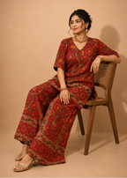 A woman sits on a wooden chair wearing a red printed kurta and wide-leg trousers set with floral motifs. The background is a solid warm beige studio backdrop.
