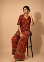 A woman models a red and gold printed two-piece kurta set while sitting on a black stool against a solid beige studio background.