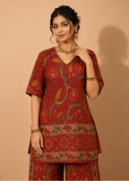 A woman models a rust-red three-piece kurta set with yellow, green, and orange floral block prints, set against a solid beige background.