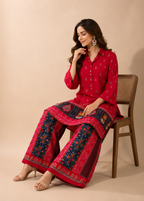 A woman seated on a wooden chair wearing a bright red traditional outfit with dark blue and red patterned wide-leg pants, against a plain beige studio background.