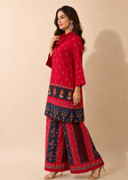 A woman modeling a red three-piece salwar kameez suit with navy blue and red printed borders, standing against a solid beige studio background.