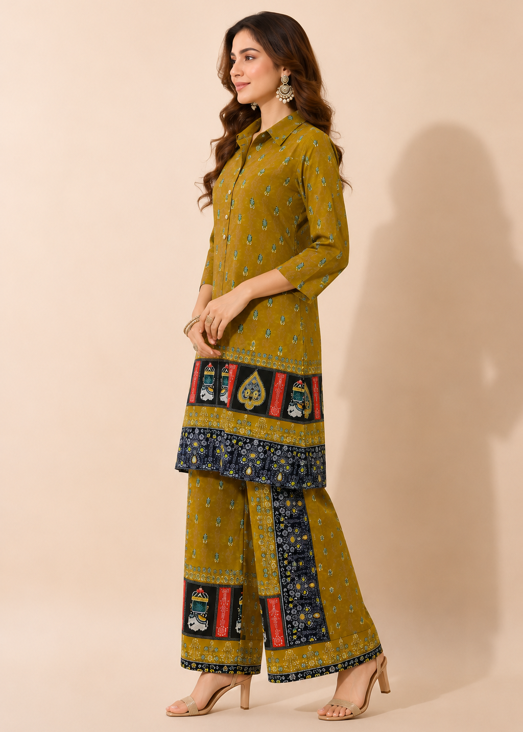 A woman modeling a mustard yellow three-piece salwar kameez suit with blue, red, and green printed borders on the kurta and wide-leg pants, set against a solid beige studio background.