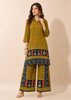 A woman models a mustard yellow three-piece salwar kameez suit set with intricate blue, red, and gold printed borders on the kurta and wide-leg trousers, set against a solid beige studio background.