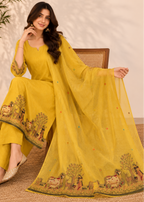 A woman models a yellow three-piece salwar kameez suit with a matching dupatta. The set features a solid yellow kurta and wide-leg trousers with detailed multicolor embroidery depicting animals and trees along the borders.