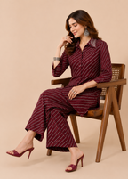 A woman seated on a wooden chair wearing a burgundy and white striped kurta set with matching wide-leg pants and heeled sandals, against a plain beige studio background.