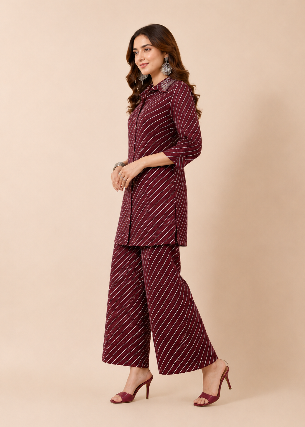 A woman modeling a matching two-piece outfit consisting of a long-sleeve button-up shirt and wide-leg trousers, both in a deep maroon color with thin white diagonal stripes, set against a solid beige studio background.