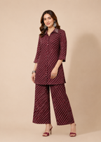 A woman modeling a matching two-piece outfit consisting of a long-sleeve button-up kurta and wide-leg trousers, both in a deep maroon color with thin white diagonal stripes, set against a solid beige studio background.