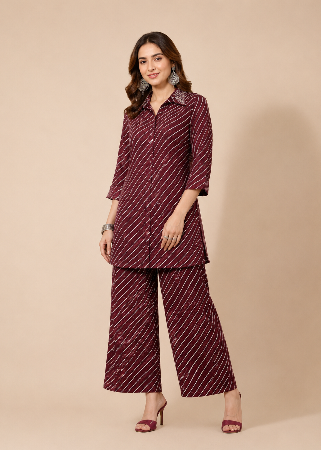 A woman modeling a matching two-piece outfit consisting of a long-sleeve button-up kurta and wide-leg trousers, both in a deep maroon color with thin white diagonal stripes, set against a solid beige studio background.