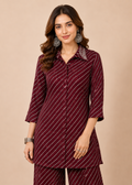 A woman modeling a matching deep maroon three-piece kurta set with thin white diagonal stripes, featuring a collared long-sleeve top, button-front styling, and coordinating trousers, set against a solid beige background.