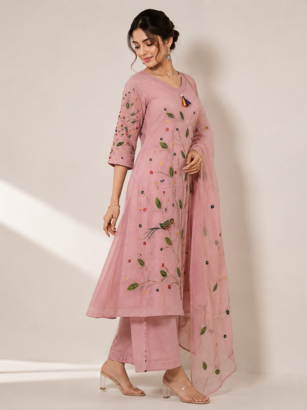 A woman models a pink traditional Indian outfit consisting of a kurta with floral embroidery, matching trousers, and a sheer dupatta, set against a plain beige studio background.