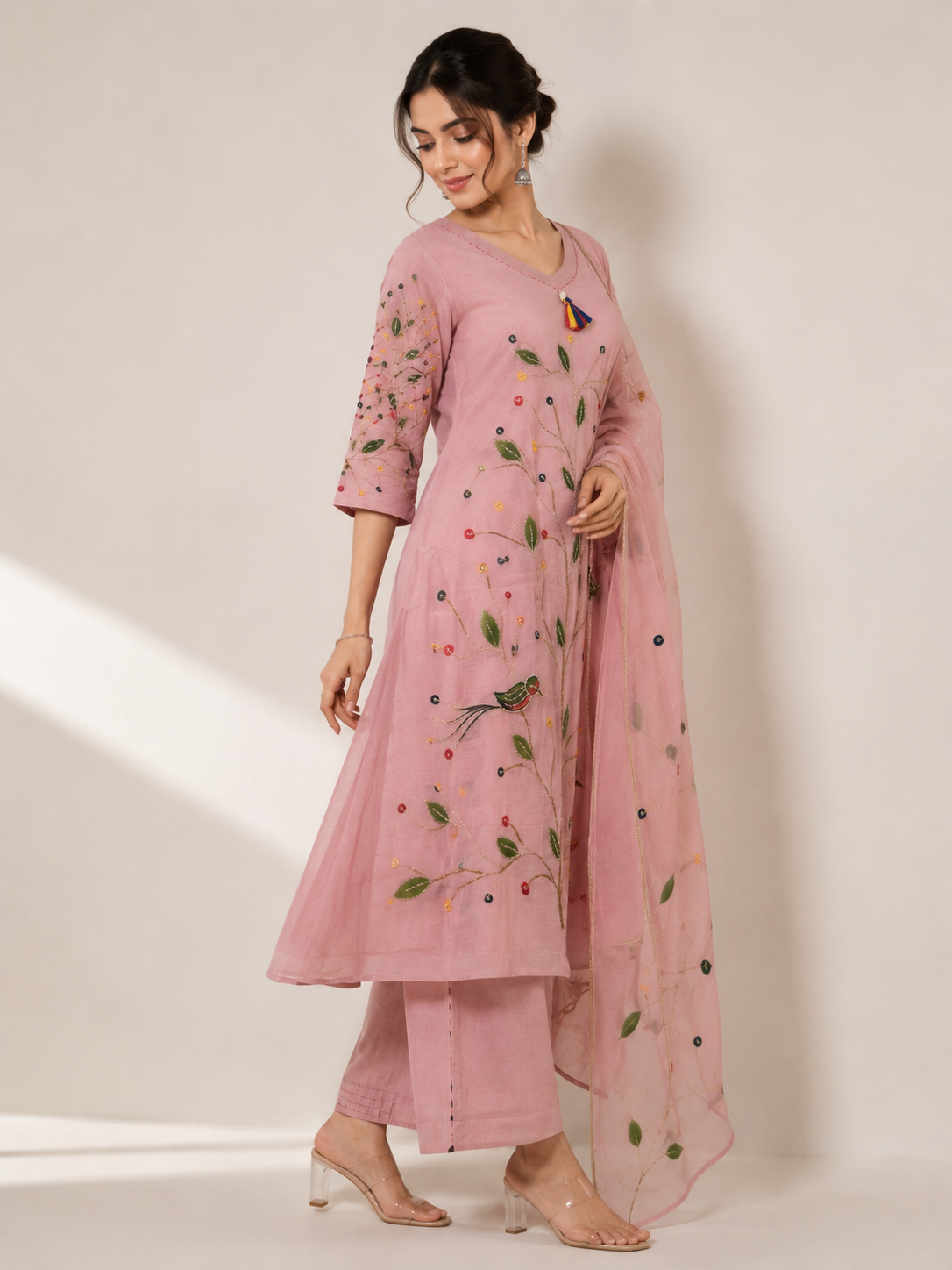 A woman models a pink traditional Indian outfit consisting of a kurta with floral embroidery, matching trousers, and a sheer dupatta, set against a plain beige studio background.