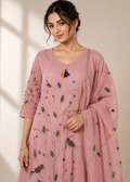A woman models a dusty pink embroidered kurta set with a matching dupatta. The kurta features delicate floral and bird embroidery in green, red, and yellow, with a colorful tassel pendant at the neckline.