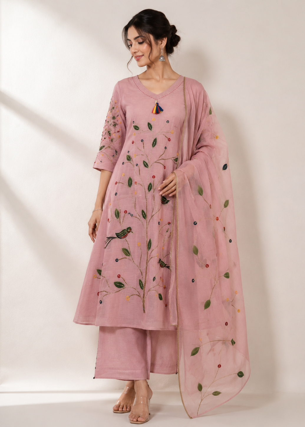 A woman models a dusty pink Indian ethnic suit set featuring a kurta with green and multicolor floral embroidery, matching trousers, and a sheer dupatta draped over her shoulder, set against a warm off-white background.
