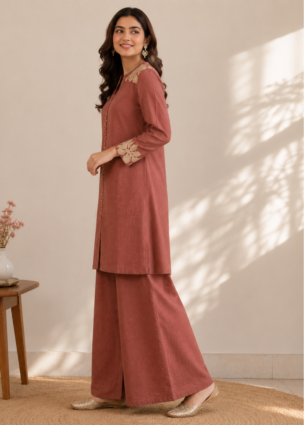A woman models a dusty rose two-piece kurta and trouser set with beige floral embroidery on the collar and cuffs, standing in a sunlit room with a small wooden side table and vase on the left.