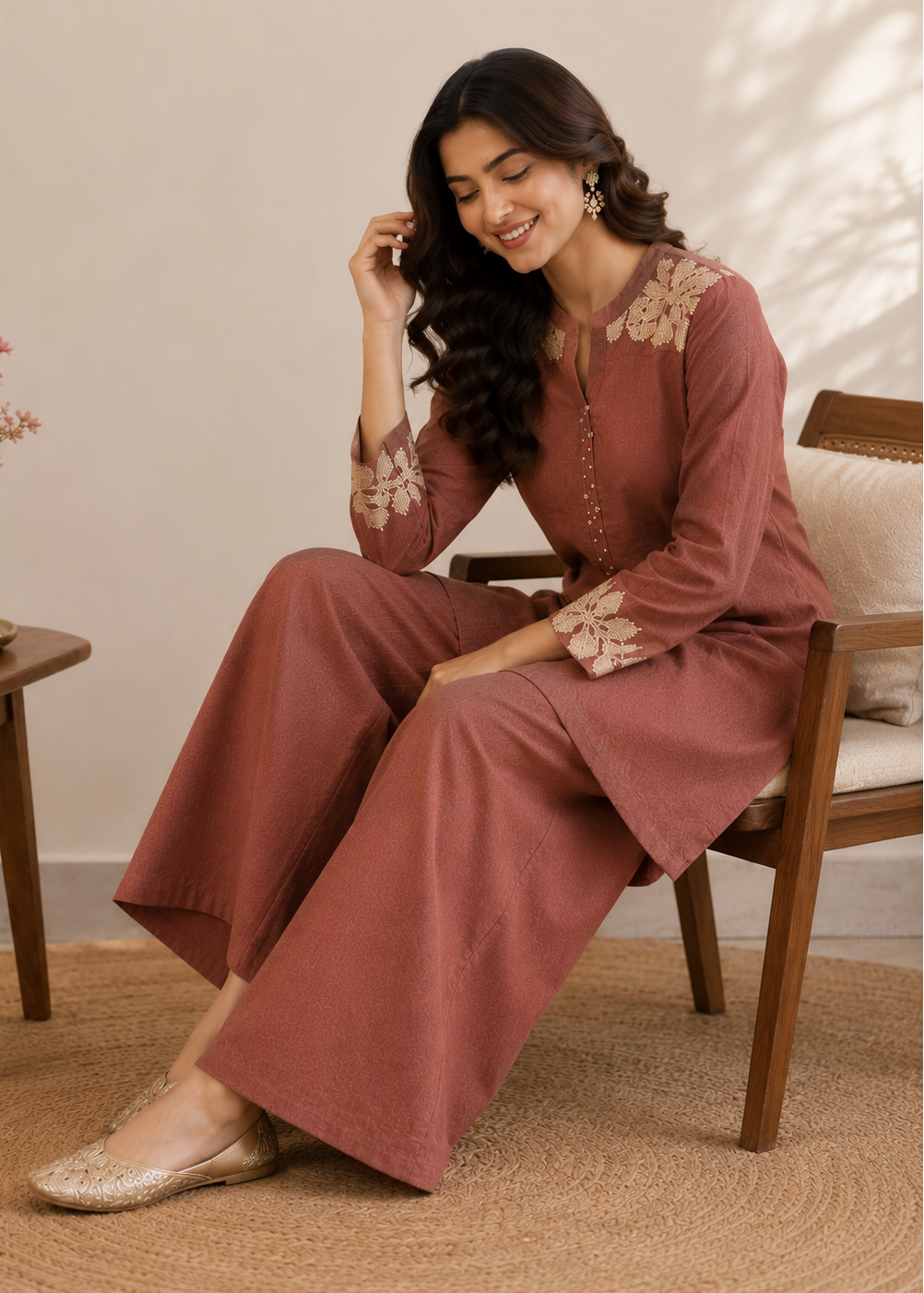 A woman wearing a dusty rose two-piece kurta set with beige floral embroidery sits on a wooden chair in a warm, neutral-toned room.