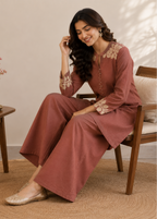 A woman wearing a dusty rose two-piece kurta set with beige floral embroidery sits on a wooden chair in a warm, neutral-toned room.