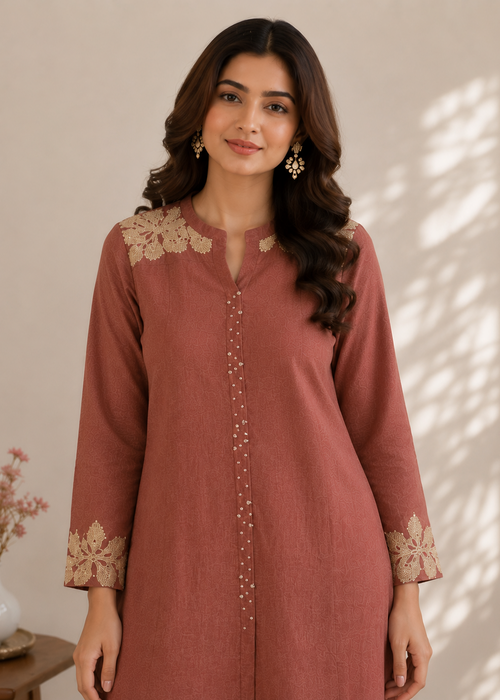 A woman wearing a dusty rose long kurta with beige floral embroidery on the shoulders and cuffs, standing against a warm beige wall with soft shadow patterns.