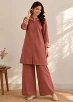 A woman models a two-piece dusty rose kurta and wide-leg trouser set with beige floral embroidery on the collar, cuffs, and button placket, set against a warm beige wall with soft leaf shadows.