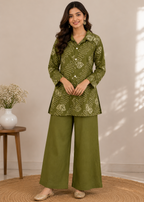 A woman models a two-piece olive green kurta set with a white abstract pattern, consisting of a long-sleeve button-up top and matching wide-leg trousers, standing on a round woven rug.