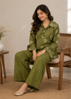 A woman sits on a wooden chair wearing an olive green two-piece pajama set with a cream-colored abstract pattern. The set includes a long-sleeve button-up shirt and matching wide-leg pants.