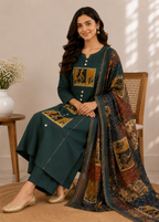 A woman seated on a wooden chair wearing a dark teal traditional outfit with ornate gold and brown embroidered panels and a matching patterned dupatta. The setting is indoors with a neutral beige wall, a small side table with a white vase of flowers, and soft shadow light across the background.