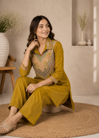 A woman sits on a woven round stool in a warm, neutral-toned room, wearing a mustard yellow kurta set with a detailed multicolor embroidered yoke. The set includes matching wide-leg trousers and a collared shirt-style top.