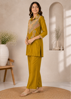 A woman models a mustard yellow two-piece kurta set with intricate multicolor embroidery on the chest, standing in a warm, neutral-toned room with decorative vases and a woven rug.