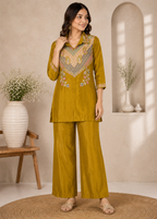 A woman models a mustard yellow two-piece kurta and trouser set with intricate multicolor embroidery on the chest, set against a warm beige interior background with decorative vases.