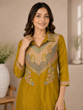 A woman wearing a mustard yellow kurta with intricate multicolor embroidery on the chest and cuffs, standing in a softly lit indoor setting with decorative vases.